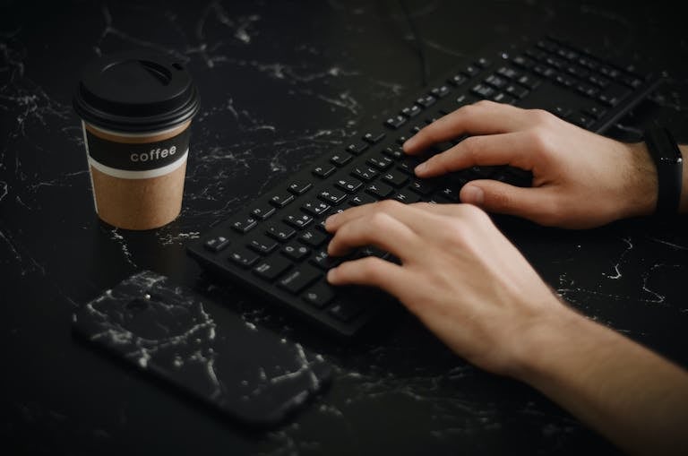 A pair of hands typing on a keyboard with a coffee cup and smartphone on a dark desk.