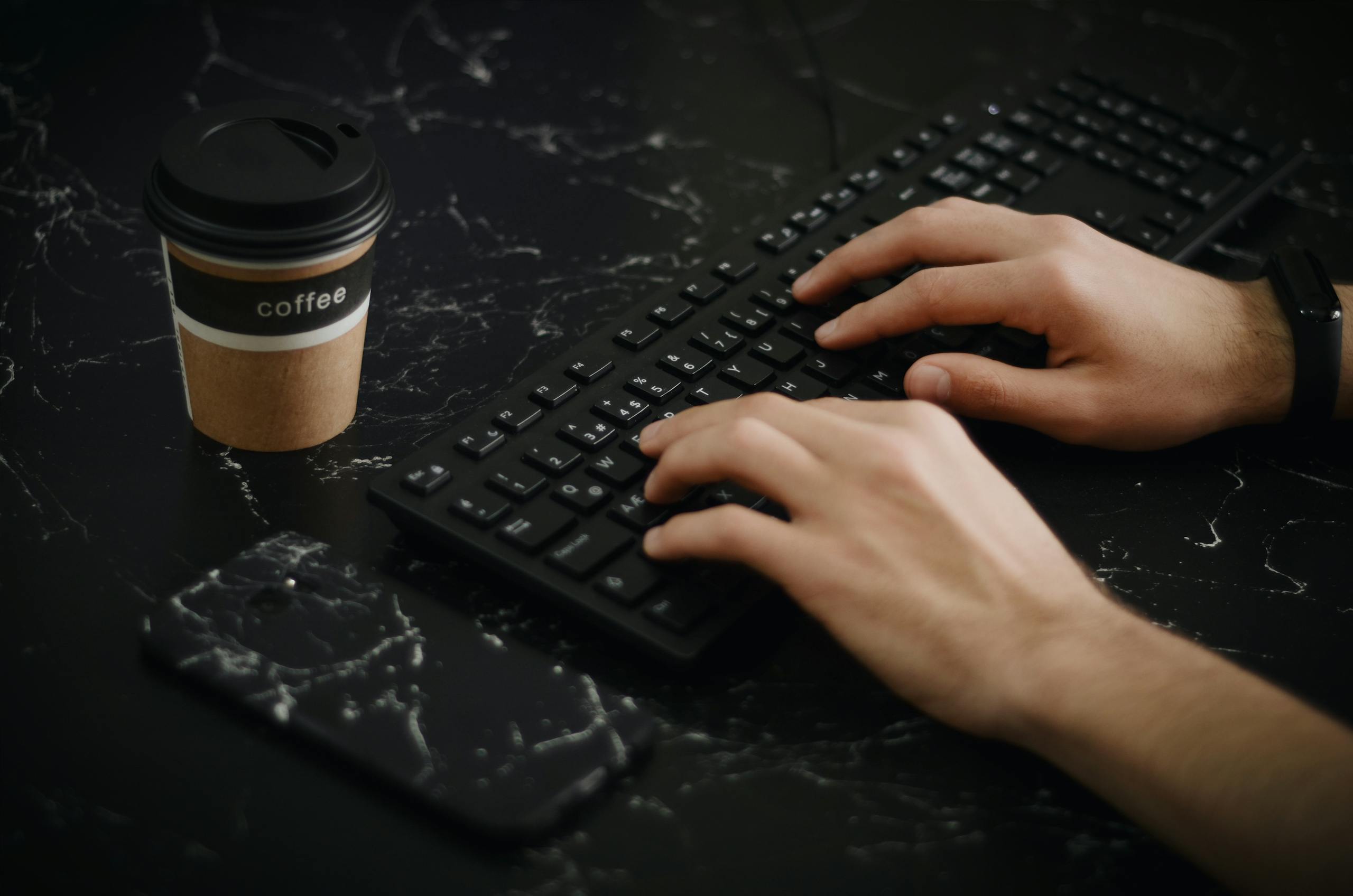 A pair of hands typing on a keyboard with a coffee cup and smartphone on a dark desk.